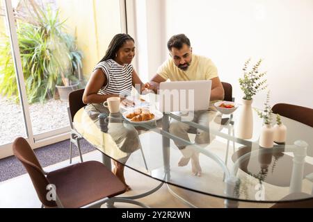 Una donna nera condivide un momento di gioia per la colazione con un uomo indiano mentre si impegna con un portatile in una luminosa e moderna sala da pranzo Foto Stock