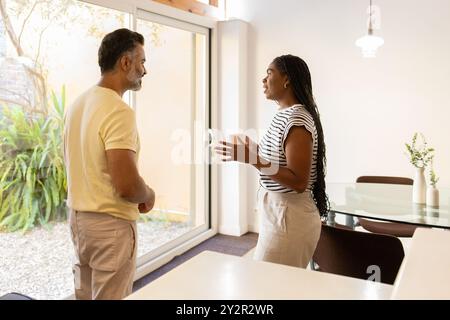 Una donna nera e un uomo indiano si impegnano in una conversazione rilassata all'interno della loro casa luminosa e moderna, in piedi vicino a una grande finestra che si affaccia su un giardino Foto Stock