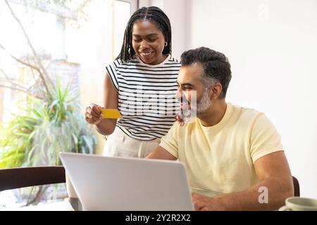 Una donna nera gioiosa che ha una carta di credito si trova accanto a un uomo indiano sorridente mentre sfogliano insieme un negozio online su un notebook al loro buio Foto Stock