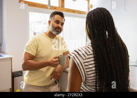 Un momento intimo in una cucina luminosa come un uomo indiano di mezza età e una donna nera si godono una conversazione mattutina con tazze di caffè Foto Stock