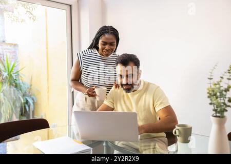 Un'immagine affascinante che cattura una donna nera gioiosa e un uomo indiano che si impegnano in un momento piacevole sopra un notebook a casa, incarnando una casa moderna che vive un Foto Stock