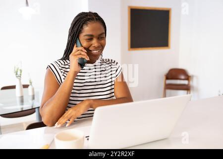 Un'allegra donna afroamericana si impegna in una conversazione telefonica mentre lavora in multitasking sul suo notebook a un tavolo da pranzo ben illuminato, raffigurando un comodo divano Foto Stock