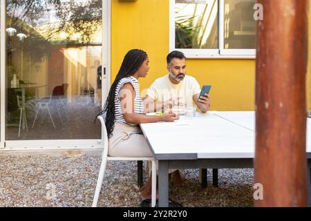 Una donna di colore e un uomo indiano siedono a un tavolo bianco all'aperto, mentre mangiano con l'uomo utilizzando uno smartphone, contro un dorso giallo brillante Foto Stock