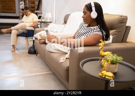 Una donna nera ascolta la musica con le cuffie mentre naviga sul suo smartphone, seduto comodamente su un divano il suo partner, un uomo, è sullo sfondo, Foto Stock