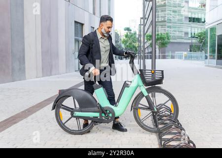 Un uomo d'affari indiano monta una bicicletta elettrica nel parcheggio di un paesaggio urbano moderno, trasudando professionalità ed ecocompatibilità. Foto Stock