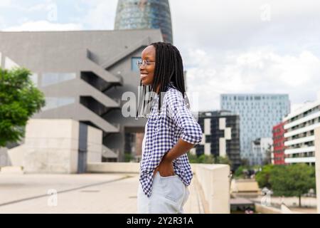 Una donna nera con i capelli intrecciati sorride mentre cammina in un moderno quartiere commerciale, mostrando fiducia e un comportamento rilassato Foto Stock