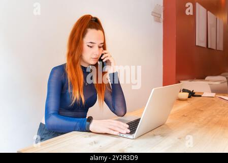 Una giovane donna dai capelli rossi si impegna in una conversazione telefonica mentre utilizza il suo notebook in uno spazio di lavoro elegante e sparso, a indicare un ci moderno Foto Stock