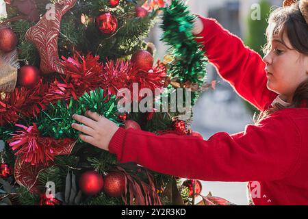 Una giovane ragazza con un maglione rosso decora un albero di Natale all'aperto, sistemando con cura la tinsel verde e rossa Foto Stock