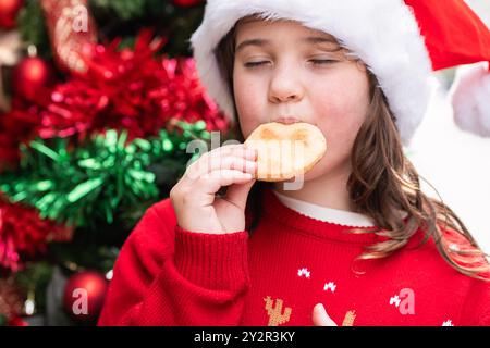 Una giovane ragazza con un cappello di Babbo Natale e un maglione rosso festoso gode di un biscotto natalizio a forma di cuore, con decorazioni colorate e luci sullo sfondo Foto Stock