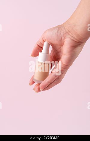 Immagine ravvicinata di una mano che stringe saldamente un flacone contagocce riempito con un composto di base beige su un morbido sfondo rosa che riflette un ap moderno Foto Stock