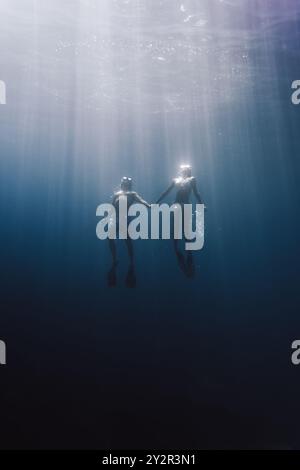 Un etereo scatto sott'acqua che cattura due subacquei, collegati da mani, immersi in un oceano sereno e blu, illuminati dalla luce naturale del sole Foto Stock