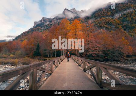 Persona che cammina su un ponte circondato da un vivace fogliame autunnale nella Valle d'Ordesa, con montagne ricoperte di nebbia sullo sfondo Foto Stock