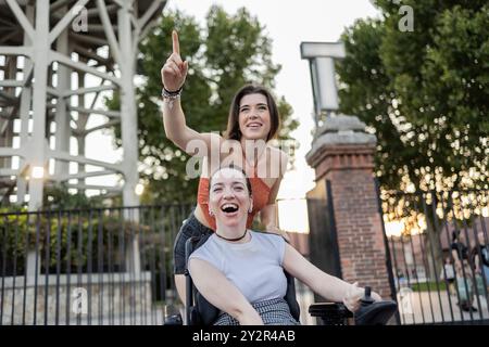 Due giovani donne vivaci che condividono un momento di gioia all'aperto; una di loro usa una sedia a rotelle che ridono insieme, simboleggiando l'amicizia e l'inclusione Foto Stock