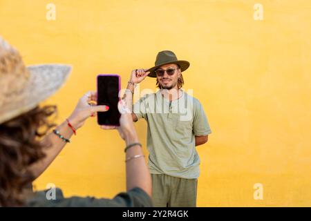 Una donna cattura una foto di un uomo gioioso con dreadlock, che indossa un cappello su un vibrante sfondo giallo. Lui non guarda scherzosamente mentre fotografa Foto Stock