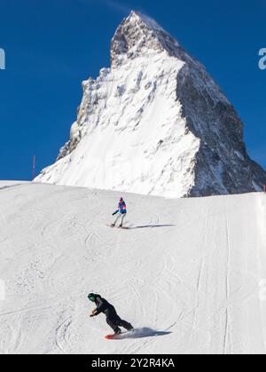Due sciatori che indossano attrezzatura e caschi scivolano lungo piste innevate incontaminate con la maestosa vetta del Cervino sullo sfondo a Zermatt, nelle Alpi svizzere. La S Foto Stock