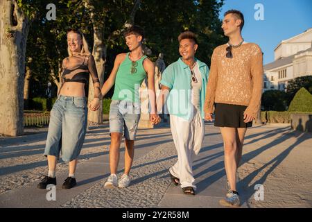 Un gruppo eterogeneo di amici LGBTQIA+ che si tengono per mano e sorridono durante una piacevole passeggiata in un parco illuminato dal sole, simboleggiando l'amicizia e l'inclusione. Foto Stock