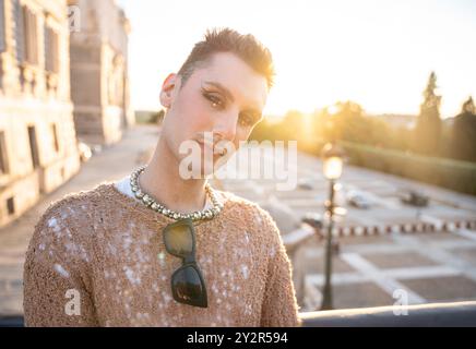 Una persona non binaria posa all'aperto con lo sfondo di un tramonto della città, mostrando sicurezza e stile Foto Stock