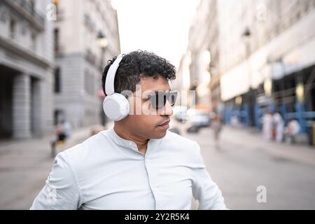 Un giovane ed elegante latino che indossa occhiali da sole e cuffie bianche percorre una vivace strada cittadina, intriso di musica. Foto Stock