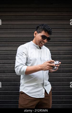 Un allegro giovane latino in un elegante abbigliamento casual ride mentre maneggia una macchina fotografica d'epoca, davanti a uno sfondo di metallo scuro. Foto Stock