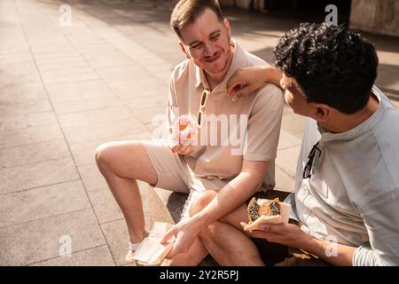 Una felice coppia gay multietnica siede su una strada cittadina, condividendo ciambelle e risate sotto la calda luce del sole, creando un momento confortevole e amorevole. Foto Stock