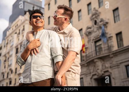 Una felice coppia gay multietnica condivide una risata mentre esplora un'area urbana storica, mostrando gesti affettuosi e attimi sportivi estivi informali Foto Stock