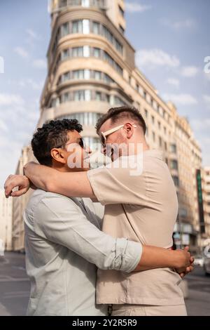 Un momento tenero tra una coppia gay multietnica viene catturato mentre si abbracciano in una vivace strada cittadina, mostrando amore e connessione in mezzo a una città Foto Stock