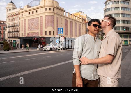 Affettuose coppie gay multietniche condividono un caldo abbraccio e bacio sulla guancia di una strada trafficata della città, con un vivace sfondo urbano caratterizzato da stile storico Foto Stock