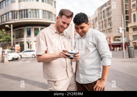 Una felice coppia gay multietnica che condivide un momento in una strada cittadina guardando le foto su una macchina fotografica d'epoca, incarnando felicità e solidarietà Foto Stock