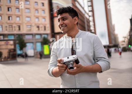 Un allegro ispanico che tiene in mano una macchina fotografica d'epoca con un sorriso, in piedi in un ambiente urbano frenetico. Lo sfondo presenta l'architettura cittadina e le passeggiate Foto Stock