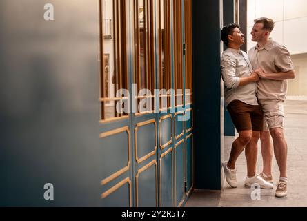 Una coppia gay multietnica condivide un momento di gioia, ridendo e abbracciando vicino a un edificio cittadino con eleganti porte blu e dorate, esprimendo amore e comicità Foto Stock