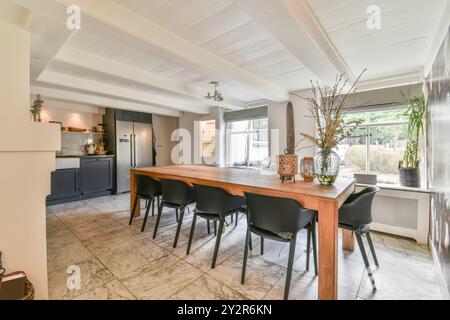 Un interno ben illuminato di una cucina moderna con una zona pranzo annessa caratterizzata da un lungo tavolo in legno, sedie nere e pavimento in marmo. Foto Stock