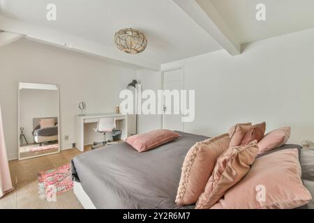 Camera moderna e ben illuminata, dotata di comodo letto con biancheria da letto grigia e rosa, grande specchio, scrivania con sedia e illuminazione decorativa. Foto Stock