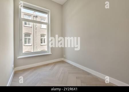 Una camera vuota presenta una grande finestra con vista di un edificio adiacente e un pavimento in legno chiaro con motivo a spina di pesce. Foto Stock