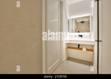 Un bagno moderno e ben illuminato caratterizzato da un elegante design minimalista con un grande specchio, illuminazione sotto gli scaffali e un ripiano in legno. Foto Stock