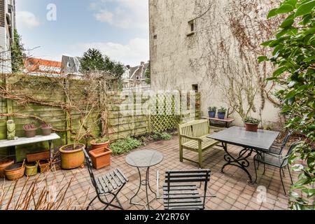 Un pittoresco giardino urbano sul retro che presenta una panchina in legno, sedie in metallo nero, un tavolo e varie piante in vaso, con uno sfondo di verde e un Foto Stock