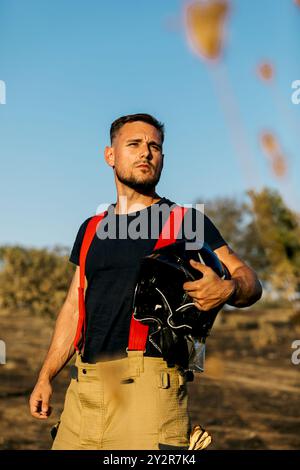 Un pompiere concentrato, in piedi in un'area boscosa, tiene un casco nero in una mano e indossa pantaloni da lavoro beige e bretelle rosse e nere Foto Stock