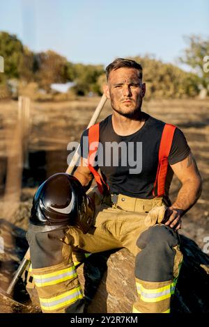 Un determinato vigile del fuoco siede tranquillo dopo aver combattuto un incendio nella foresta, tenendo in mano un casco e appoggiandosi su una roccia nel mezzo di un paesaggio carbonizzato Foto Stock