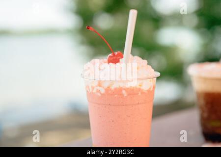 Primo piano di un frullato di fragole ricoperto di panna montata e ciliegia, con sfondo esterno sfocato. Foto Stock