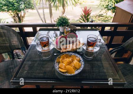 Una deliziosa colazione tropicale con mango fresco, pasticcini e tè viene servita su un balcone. L'ambiente e' luminoso e invitante, circondato da vib Foto Stock