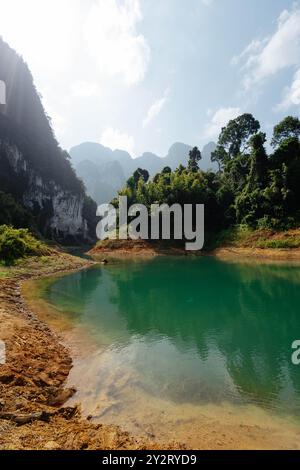 Un tranquillo lago turchese riflette la vegetazione lussureggiante circostante e le torreggianti montagne sotto un cielo luminoso. Questo idilliaco ambiente naturale invita a Foto Stock