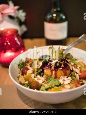Primo piano di un'insalata gourmet con verdure miste, pomodori ciliegini, noci e una palla di formaggio condita con salsa di frutti di bosco in una ciotola bianca Foto Stock
