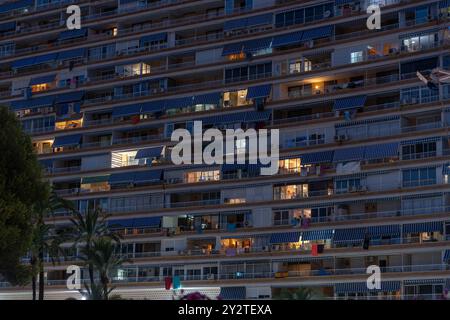 Un edificio residenziale ad Alicante, in Spagna, con tende a strisce blu e finestre illuminate durante la notte, creando una tranquilla scena urbana. Foto Stock