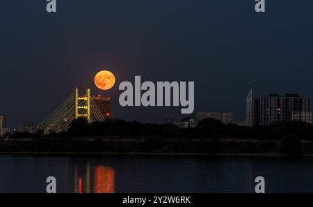 Una splendida vista notturna di un paesaggio urbano con una luna piena che si innalza dietro un ponte, che si riflette su un fiume tranquillo. Foto Stock