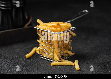 Una porzione di patatine fritte dorate in un piccolo cestino di metallo su sfondo scuro, con alcune patatine sparsi intorno. Foto Stock