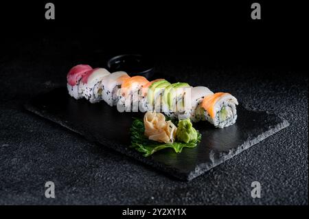 Un piatto di sushi ben organizzato con vari tipi di sushi roll, guarnito con wasabi e zenzero su un piatto scuro su sfondo nero. Foto Stock