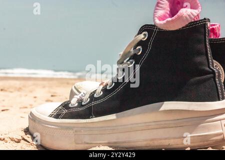 Le sneaker Converse da donna in bianco e nero con farfalle rosa si stagliano su una spiaggia sabbiosa. Vacanze al mare in estate. Cartolina estiva vintage Foto Stock