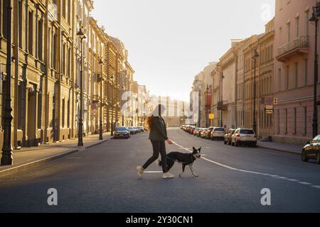 Una donna attraversa la strada con il suo cane collie al sorgere del sole, con una luce soffusa che illumina gli edifici. L'immagine cattura un momento tranquillo Foto Stock