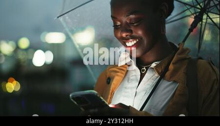 Outdoor, donna nera e ombrello con smartphone di notte, messaggi di testo e chat online sui social media. Città, donna e cellulare con pioggia Foto Stock