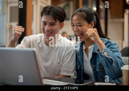 Due giovani amici asiatici allegri ed eccitati festeggiano insieme buone notizie, guardando uno schermo di un notebook con espressioni entusiastiche e mostrando il Foto Stock