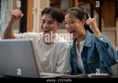 Due giovani amici asiatici allegri ed eccitati festeggiano insieme buone notizie, guardando uno schermo di un notebook con espressioni entusiastiche e mostrando il Foto Stock